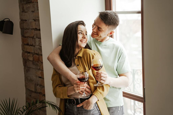 Bella coppia che si abbraccia con bicchieri di vino a casa, vibrazioni romantiche. Felice giovane famiglia si trasferì in un nuovo appartamento - Foto, immagini