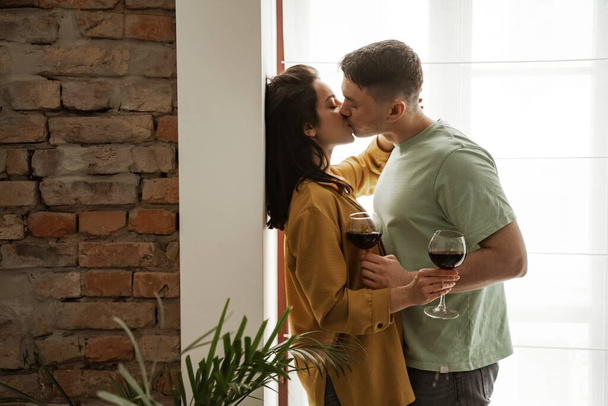 Preciosa pareja con copas de vino, besos en casa, vibraciones románticas. Familia joven feliz se mudó a nuevo apartamento - Foto, Imagen