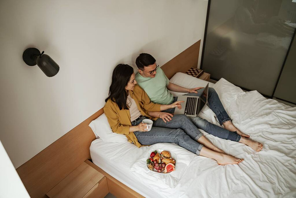 Lovely couple watching movie with romantic feelings, sitting on bed with coffee and plate of croissant and fresh fruits. Happy family moving to new apartment - Photo, Image
