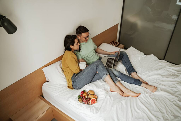 Lovely couple watching movie with romantic feelings, sitting on bed with coffee and plate of croissant and fresh fruits. Happy family moving to new apartment - Foto, imagen