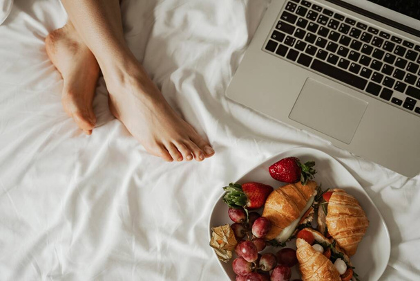 Close up plate of croissant, strawberry and fruits with red wine and laptop in bed. Happy family moving to new apartment - Fotoğraf, Görsel