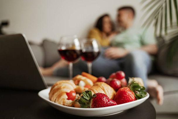 Close up plate of croissant and strawberry, lovely couple kissing with romantic feeling sitting on couch in living room. Happy family moving to new apartment - Photo, Image