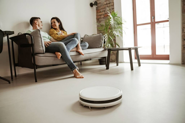 Lovely young couple showing sitting on couch in living room while robot vacuum cleaner working. Happy family moving to new apartment - Foto, afbeelding