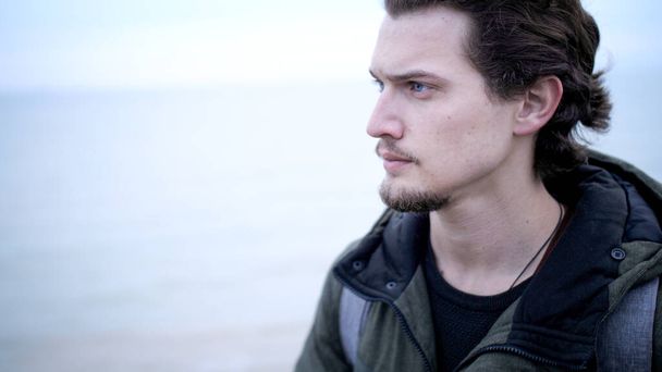 Attraktiver blauäugiger junger Mann mit Bart und lockigem Haar einsames Porträt an der Küste - Foto, Bild