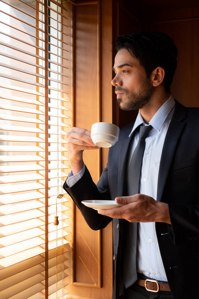 πορτρέτο του νεαρού όμορφου επιχειρηματία της Μέσης Ανατολής που φοράει κοστούμι και γραβάτα, πίνοντας καφέ ενώ κάνει ένα διάλειμμα στο γραφείο. επιχειρηματική ιδέα - Φωτογραφία, εικόνα