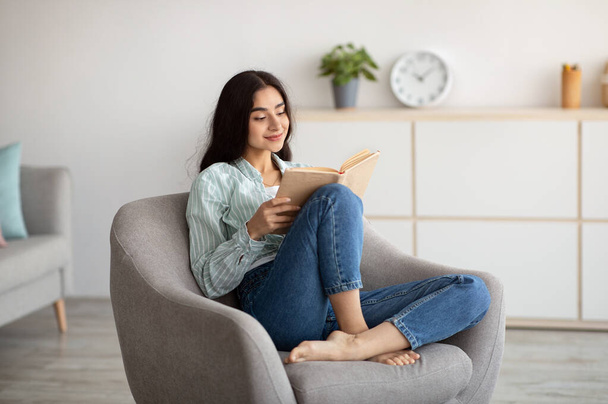 Stay home pastimes. Millennial Indian woman sitting in cozy armchair with open book indoors - Фото, изображение