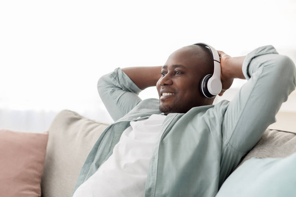 Afrika kökenli orta yaşlı, kablosuz kulaklıklı adam oturma odasındaki kanepede müzik dinliyor, uzayı kopyalıyor. - Fotoğraf, Görsel