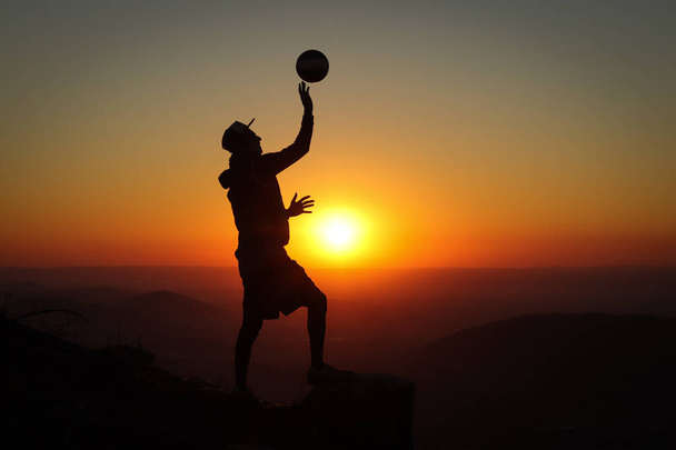 Profesyonel voleybol oyuncusu bir dağın tepesinde bir kütük üzerinde duruyor ve gün batımında bir elinde voleybol topuyla oynuyor. Geceleri siluet. - Fotoğraf, Görsel