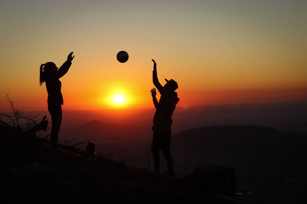 Νεαρό ζευγάρι βόλεϊ απολαμβάνοντας την κορυφή ενός ροζ βουνού με μια μπάλα στο ηλιοβασίλεμα. Οι σιλουέτες μιας γυναίκας και ενός παίκτη να τσιμπάνε τα δάχτυλά τους με μια μπάλα. Στύλος βόλεϊ, χτύπημα. Υπέρβαση. - Φωτογραφία, εικόνα
