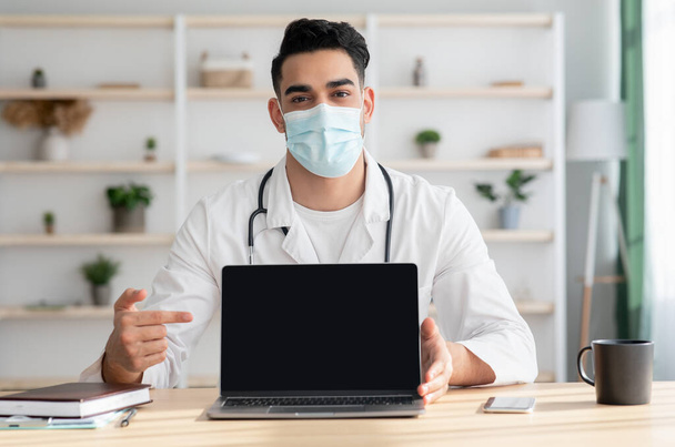 Middle-eastern man doctor in face mask pointing at laptop - Photo, Image