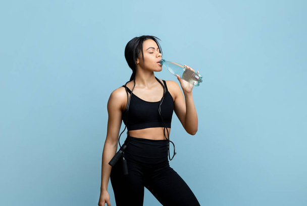 Sports hydration concept. Sporty african american woman drinking mineral water from bottle at blue studio background - Foto, Imagem