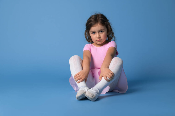 Konzentrierte bezaubernde Kinderballerina, die ihre Beine streckt, bevor sie Ballett tanzt. Porträt auf blauem Hintergrund mit Platz für Text - Foto, Bild