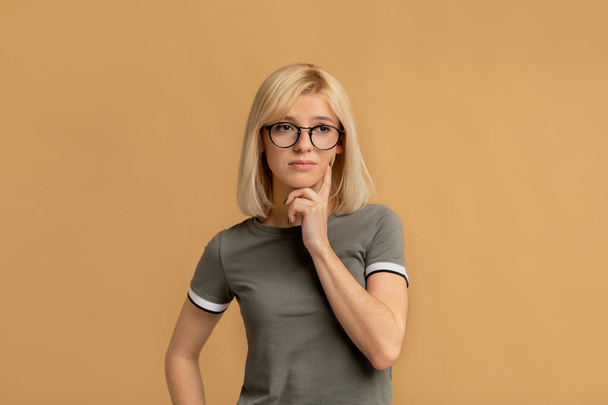 Pensive woman in glasses thinking about question, touching chin and looking away, beige background - Photo, Image