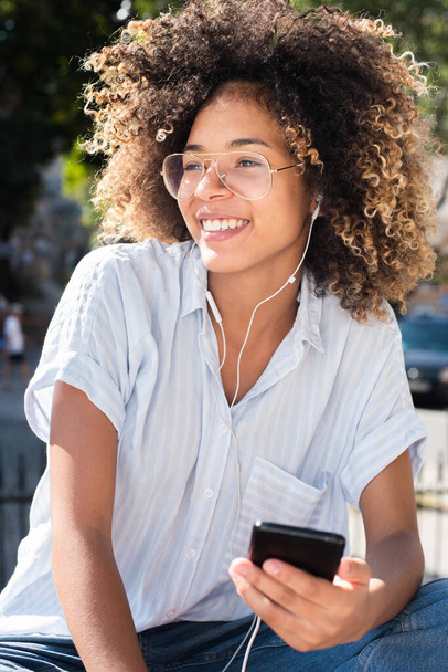 肖像画笑顔若いアフリカ系アメリカ人女性が携帯電話やイヤフォンで外に座って - 写真・画像