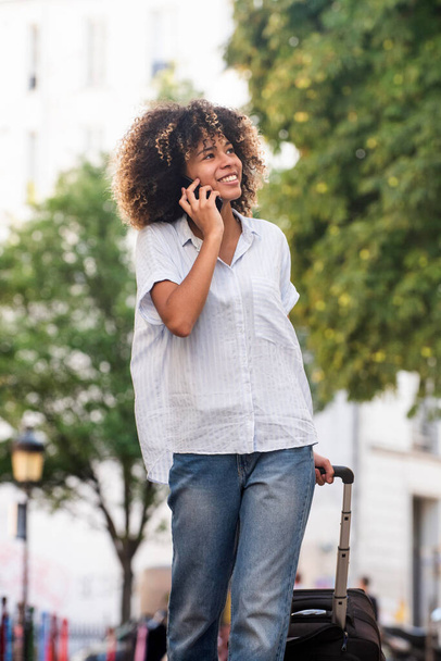 Portret gelukkig jong Afrikaans Amerikaans reizen vrouw lopen en praten met mobiele telefoon en koffer in de hand - Foto, afbeelding