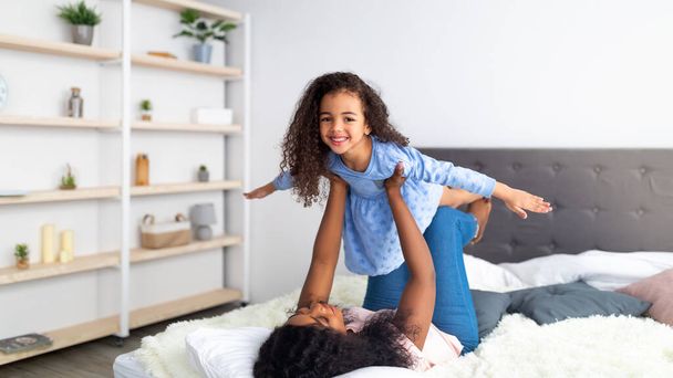 Glückliche schwarze Mutter und ihre süße Tochter beim Flugzeugspiel auf dem Bett zu Hause, Panorama. Familienbande - Foto, Bild
