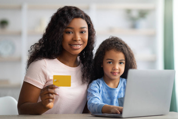 Online winkelen. Gelukkig zwart vrouw en haar dochter met behulp van credit card en laptop om goederen te kopen op het web van thuis - Foto, afbeelding