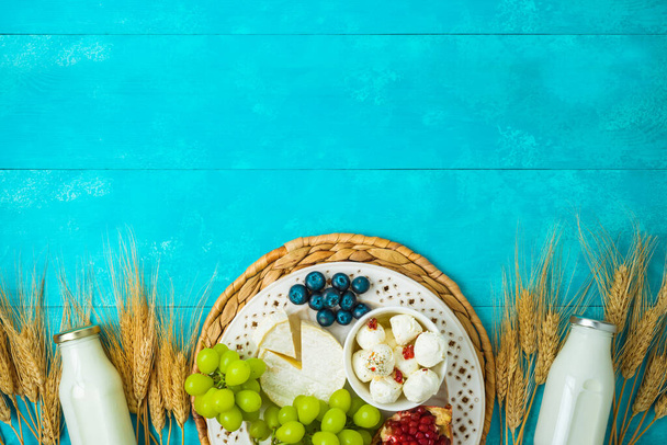 Jewish holiday Shavuot celebration concept with cheese, milk bottle, wheat ears and fruits on wooden blue table background - 写真・画像