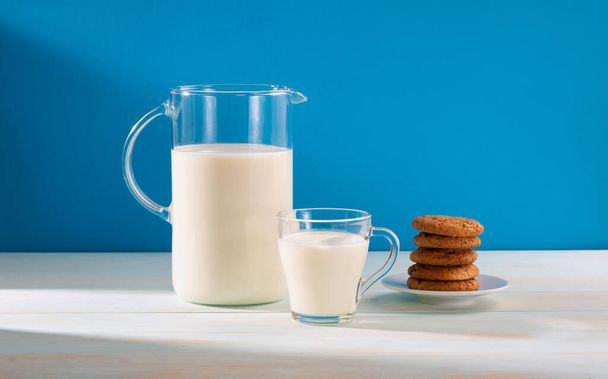 Dzbanek i szklanka mleka z ciasteczkami na drewnianym stole, na niebieskim tle.Kopiuj przestrzeń. - Zdjęcie, obraz