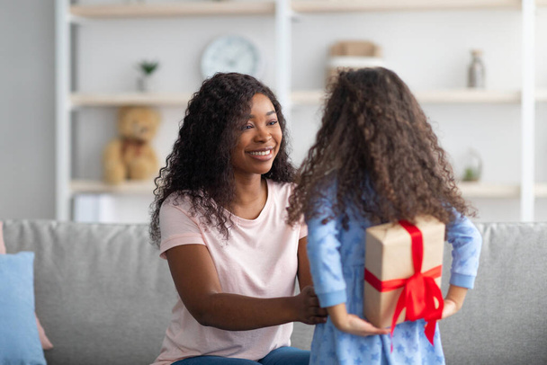 Klein zwart meisje verbergen verpakt geschenkdoos voor vakantie achter haar rug, gefeliciteerd haar moeder met verjaardag thuis - Foto, afbeelding