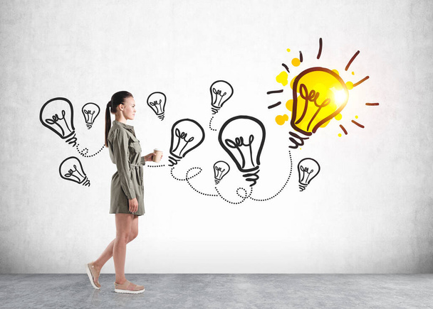 European attractive young woman in casual dress holding to go coffee cup and standing near wall with colorful business idea lightbulb sketch. Concept of creativity and brainstorming - Φωτογραφία, εικόνα