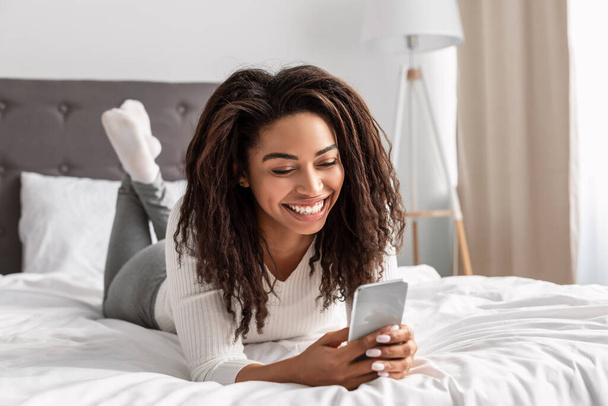 Happy black woman lying in bed reading text message - Foto, imagen