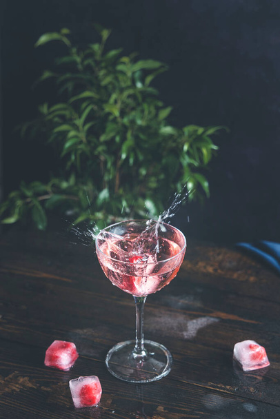 Un soupçon de champagne rose rose dans un verre élégant avec des framboises congelées dans un glaçon sur une surface de table en bois sombre, à proximité, peu profonde du champ - Photo, image