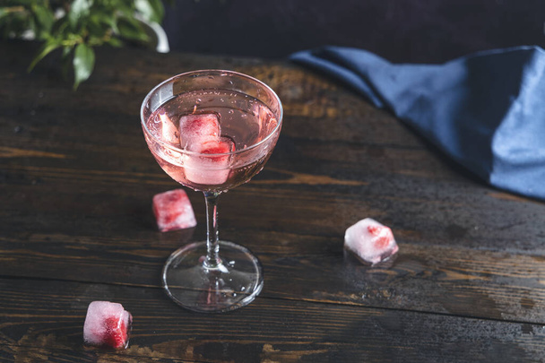 Copo elegante de champanhe rosa rosa com framboesas congeladas em cubo de gelo e gotas de água na superfície da mesa de madeira escura, close up, profundidade rasa do campo - Foto, Imagem