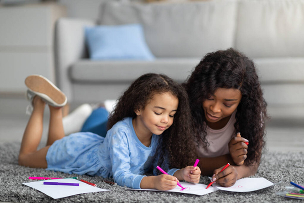 Affectionate zwart mam tekening met haar dochtertje op vloer in huis - Foto, afbeelding