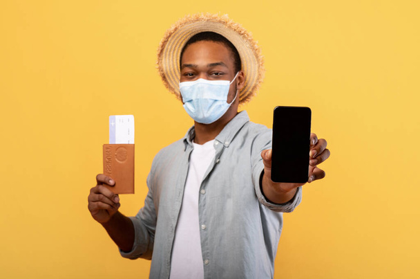 Jonge zwarte man in beschermend masker met paspoort, tickets en mobiele telefoon met mockup voor mobiele app of website ontwerp - Foto, afbeelding