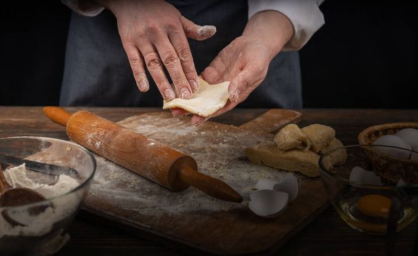 Women's hands, flour and dough. A woman, in an apron cooking dough for homemade baking, a rustic home cozy atmosphere, a dark background with unusual lighting. - Photo, Image