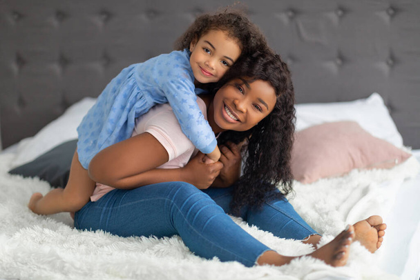 Mutlu siyah anne ve küçük kızı gündelik kıyafetleriyle yatakta sarılıyorlar. - Fotoğraf, Görsel