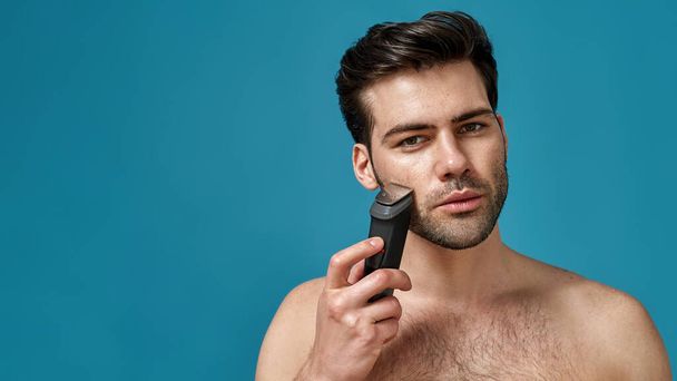 Beauty portrait of handsome young man using electric shaver isolated over blue background - Foto, Bild