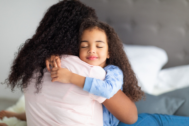 Rakastava pieni musta tyttö rento kuluminen halaa äitiään sängyssä kotona. Vahva perhesuhteiden käsite - Valokuva, kuva