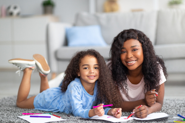 Θετική μαύρη μαμά και η ευτυχισμένη κόρη της ζωγραφίζουν μαζί στο πάτωμα στο σπίτι. Εννοιολογική έννοια του γονικού παιδιού - Φωτογραφία, εικόνα