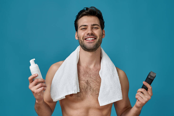 Attractive naked guy with white towel around his neck smiling at camera, holding a shaving foam and electric shaver, posing isolated over blue background - Photo, Image