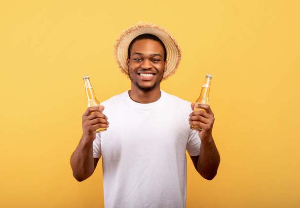 Iloinen nuori afroamerikkalainen mies, jolla on kaksi pulloa olutta ja hymyilee kameralle keltaisella studiotaustalla - Valokuva, kuva