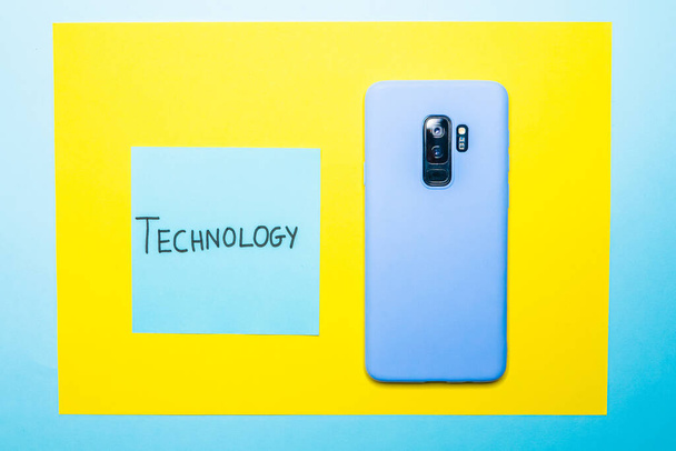A closeup shot of a mobile phone and a sticky note with "Technology" writing on a yellow surface - Zdjęcie, obraz