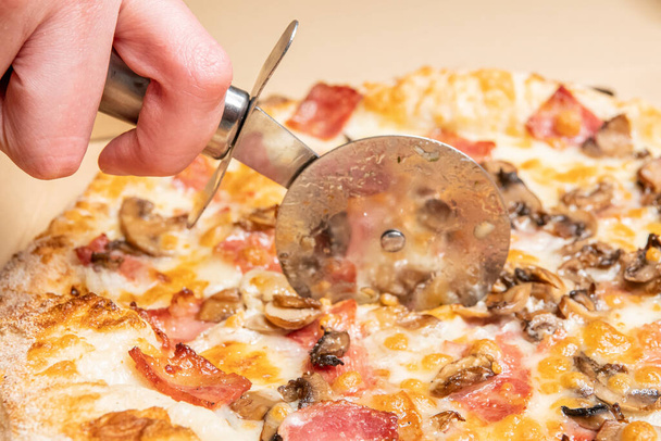 A closeup of a person cutting a delicious pizza with bacon, cheese, mushrooms, and sauces with a wheel - Photo, Image