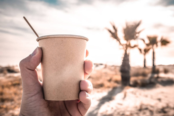 A hand holding a coffee with milk on a brown disposable paper cup against cloudy sky - Fotoğraf, Görsel