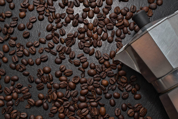A top view of the Moka pot on the black surface covered with scattered roasted coffee beans - Photo, image