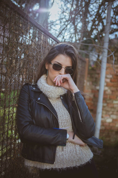 A confident brunette woman with sunglasses leaning against a metal fence wearing a leather jacket - Fotografie, Obrázek