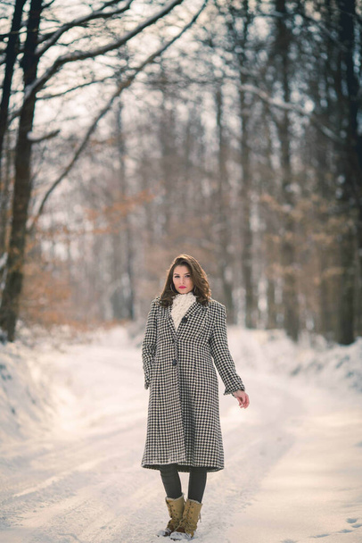 A full body shot of a young brunette girl in a stylish winter outfit walking down the forest path in winter - Zdjęcie, obraz