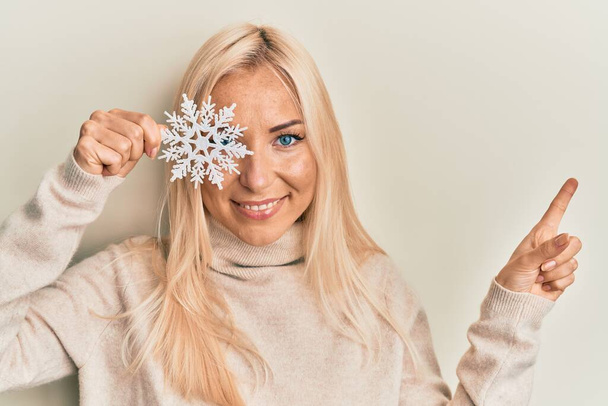 Giovane donna bionda che tiene il fiocco di neve sopra l'occhio sorridente felice indicando con mano e dito di lato  - Foto, immagini