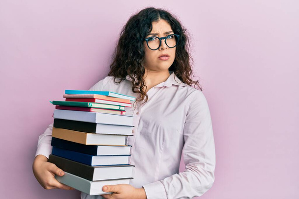Mladá brunetka s kudrnatými vlasy drží hromadu knih v šoku tvář, vypadá skeptický a sarkastický, překvapený s otevřenými ústy  - Fotografie, Obrázek