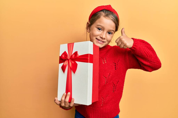 Malá krásná dívka drží dárek s úsměvem šťastný a pozitivní, palec nahoru dělá vynikající a schvalovací znamení  - Fotografie, Obrázek