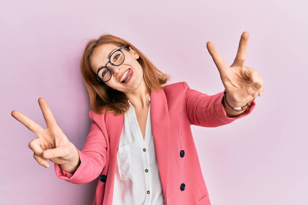 Jeune femme caucasienne portant un style d'affaires et des lunettes souriant avec la langue montrant les doigts des deux mains faisant signe de victoire. numéro deux.  - Photo, image