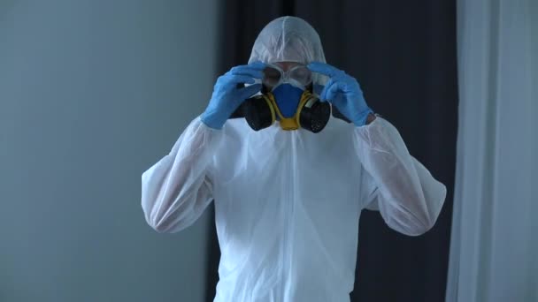 Egy férfi védőruhát, védőszemüveget és orvosi maszkot visel, otthon ül a kanapén, és kesztyűt vesz fel. A coronavirus covid-19 világjárvány elleni védekezési intézkedések. - Felvétel, videó
