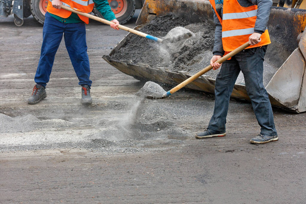 Dopravní dělníci v jasně oranžových reflexních vestách po odstranění starého asfaltu ze silnice odstraňují silniční trosky do kbelíku. Kopírovat prostor. - Fotografie, Obrázek