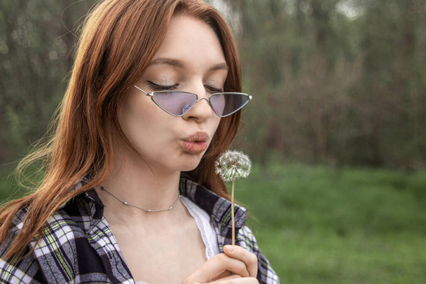 Girl blowing on a dandelion on the background of nature. Portrait of a girl with red hair - Photo, Image